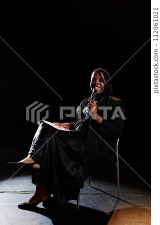 Vertical full length portrait of smiling young woman speaking to microphone on stage while sitting in chair with spotlight 112961021