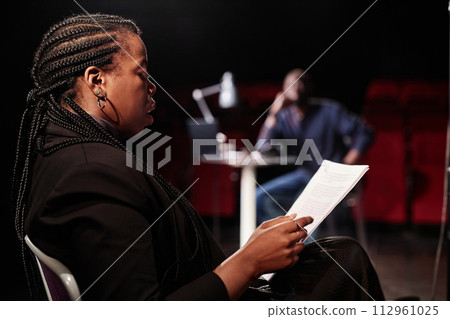 Side view portrait of African American woman reading script while sitting in chair on stage with low lighting copy space Side view portrait of African American woman reading script while sitting in chair on stage with low lighting copy space 112961025