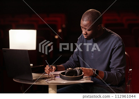 Portrait of Black adult man as theater director taking notes sitting at table with laptop copy space Portrait of Black adult man as theater director taking notes sitting at table with laptop copy space 112961044