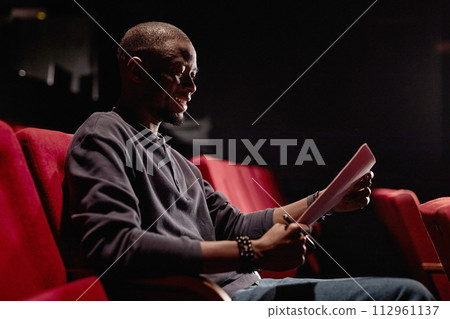 Side view portrait of smiling Black man sitting in audience and reading script in low light at theater copy space 112961137