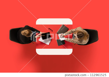 Top view. Two young business ladies sitting at common table with documents and discussing work issues against red background Top view. Two young business ladies sitting at common table with documents and discussing work issues against red background 112961525