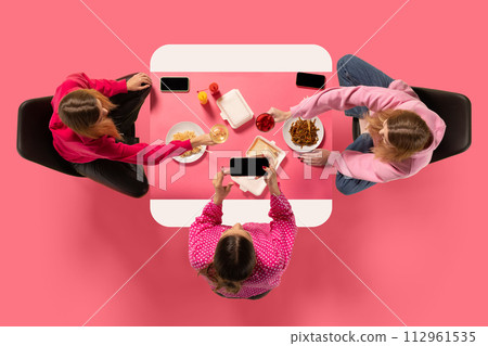 Creative collage. Top view portrait of two young girl sitting at table eating and speaking against pink background. Creative collage. Top view portrait of two young girl sitting at table eating and speaking against pink background. 112961535