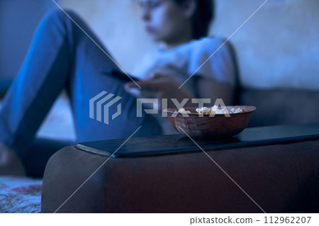 Close up view of blurred woman sitting on sofa with remote control watching TV. Selective focus on plate with popcorn. Concept of insomnia, addiction, depression, problems. Background with copy space. 112962207