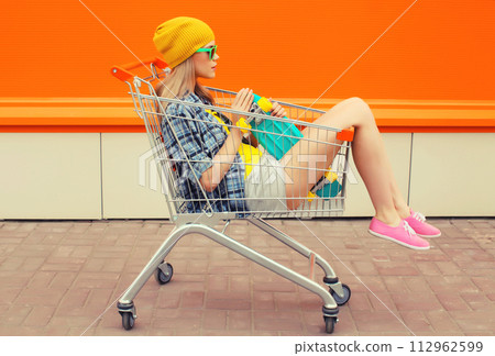 Stylish modern young woman sitting in shopping trolley cart in colorful clothes on city street 112962599
