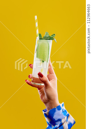 Female hand holding glass of gin tonic with cucumber and striped straw against vivid yellow studio background. 112964663
