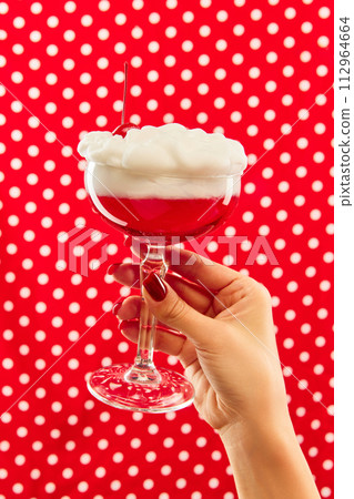 Sweet and sour. Hand holding cocktail glass decorated with cherry against red background with white polka dot. Sweet and sour. Hand holding cocktail glass decorated with cherry against red background with white polka dot. 112964664