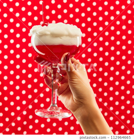 Sweet and sour. Hand holding cocktail glass decorated with cherry against red background with white polka dot. Sweet and sour. Hand holding cocktail glass decorated with cherry against red background with white polka dot. 112964665