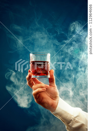 Male hand holds glass of whiskey, rum in smoke against gradient blue background with negative space to insert text. Male hand holds glass of whiskey, rum in smoke against gradient blue background with negative space to insert text. 112964666