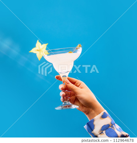 Unrecognizable woman holding glass of sweet and sour cocktail decorated with slice of carambola against blue studio background. Unrecognizable woman holding glass of sweet and sour cocktail decorated with slice of carambola against blue studio background. 112964678