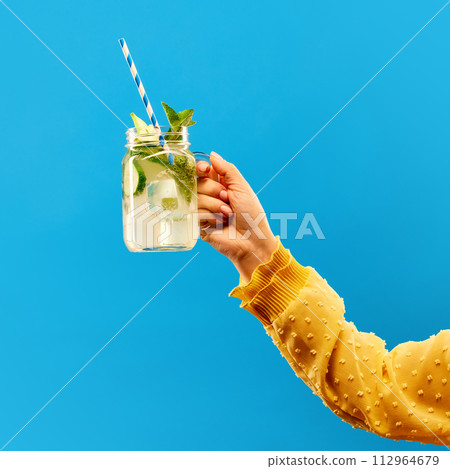 Female hand holding glass of cold refreshing citrus lemonade, mojito with straw against blue background. 112964679
