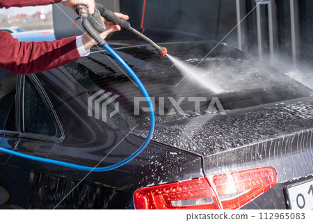 A man washes off the foam while washing the car. A vehicle is engulfed in soapy foam during a car wash, covering the tires, wheels, hood, and entire exterior of the car. Self-service car wash 112965083