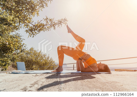 Fitness woman sea. Happy middle aged woman in orange sportswear exercises morning outdoors on yoga mat with laptop in park over ocean beach. Female fitness pilates yoga routine. Healthy lifestyle. 112966164