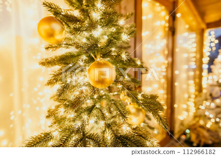 A Christmas tree with gold ornaments and lights. The tree is decorated with gold balls and is lit up with lights. The tree is placed in a room with a window A Christmas tree with gold ornaments and lights. The tree is decorated with gold balls and is lit up with lights. The tree is placed in a room with a window 112966182