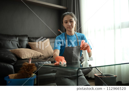 Millennial woman wearing gloves wiping dust and cleaning glass table in living room Millennial woman wearing gloves wiping dust and cleaning glass table in living room 112966520
