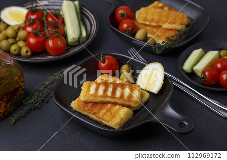 Grilled halloumi cheese in a black pan with olives, tomatoes, cucumbers, thyme and lemon. Traditional cheese of the Eastern Mediterranean. Black background, selective focus, close-up. Grilled halloumi cheese in a black pan with olives, tomatoes, cucumbers, thyme and lemon. Traditional cheese of the Eastern Mediterranean. Black background, selective focus, close-up. 112969172