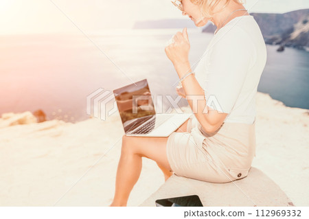Digital nomad, Business woman working on laptop by the sea. Pretty lady typing on computer by the sea at sunset, makes a business transaction online from a distance. Freelance, remote work on vacation 112969332