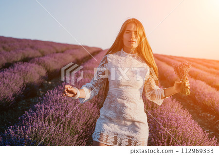 Woman lavender field. Happy carefree woman in a white dress walking in a lavender field and smelling a lavender bouquet on sunset. Ideal for warm and inspirational concepts in wanderlust and travel. 112969333