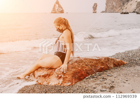 Woman travel portrait. Happy woman with long hair poses on a red volcanic rock at the beach. Close up portrait cute woman in black bikini, smiles at the camera, with the sea in the background. Woman travel portrait. Happy woman with long hair poses on a red volcanic rock at the beach. Close up portrait cute woman in black bikini, smiles at the camera, with the sea in the background. 112969339