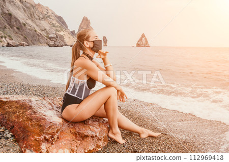 Woman travel portrait. Happy woman with long hair poses on a red volcanic rock at the beach. Close up portrait cute woman in black bikini, smiles at the camera, with the sea in the background. Woman travel portrait. Happy woman with long hair poses on a red volcanic rock at the beach. Close up portrait cute woman in black bikini, smiles at the camera, with the sea in the background. 112969418