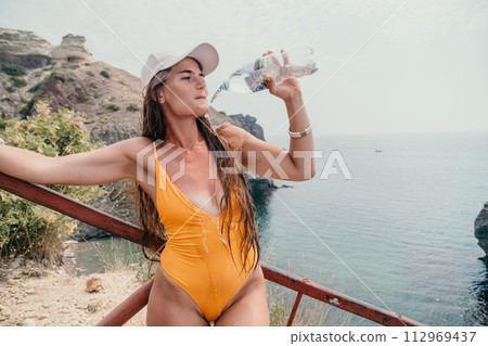 Woman travel sea. Happy tourist enjoy taking picture outdoors for memories. Woman traveler drinks water at the edge of the cliff on the sea bay of mountains, sharing travel adventure journey 112969437