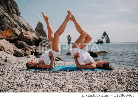 Woman sea yoga. Two Happy women meditating in yoga pose on the beach, ocean and rock mountains. Motivation and inspirational fit and exercising. Healthy lifestyle outdoors in nature, fitness concept. 112969463