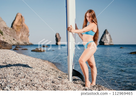Woman sea sup. Close up portrait of happy young caucasian woman with long hair looking at camera and smiling. Cute woman portrait in a blue bikini posing on sup board in the sea Woman sea sup. Close up portrait of happy young caucasian woman with long hair looking at camera and smiling. Cute woman portrait in a blue bikini posing on sup board in the sea 112969465