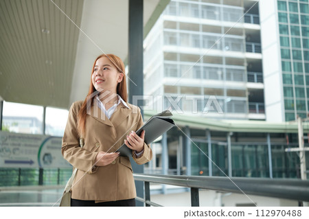 A young Asian businesswomen wearing a suit holding files standing in a big city on a busy downtown street. Young Asian businesswoman using smartphone texting to contact clients. 112970488