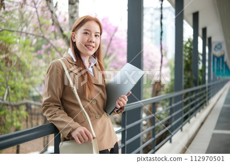 A young Asian businesswomen wearing a suit holding files standing in a big city on a busy downtown street. Young Asian businesswoman using smartphone texting to contact clients. 112970501