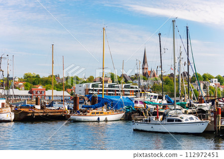 Scenic view fishing boats vessel docked Flensburg marina harbor, architectural silhouettes church cityscape background. Moored sailing vessel fjord harbour port Schleswig-Holstein Northern Germany 112972451