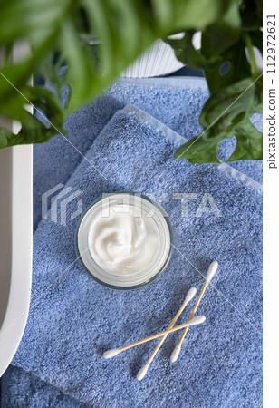 Opened cream jar on blue towels near basin and green monstera in bath, beauty routine 112972621