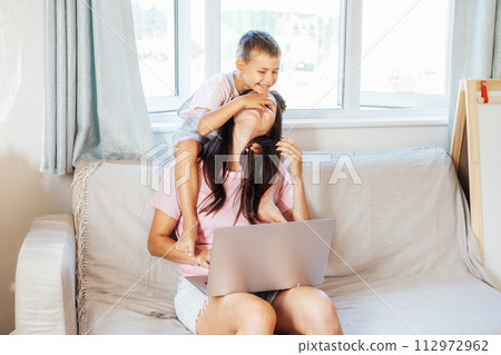 Young mother trying to work on laptop at home while her son wants to play. Childcare and working mom concept. Child make noise and disturb a woman at work. Homeschooling and freelance work. Young mother trying to work on laptop at home while her son wants to play. Childcare and working mom concept. Child make noise and disturb a woman at work. Homeschooling and freelance work. 112972962