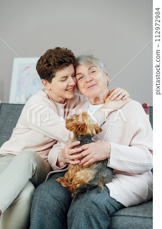 Portrait of happy senior 70s mother, adult neutral gender daughter and Yorkshire terriers dog hugging. Happy family enjoying weekend together in living room. Spending quality time in retiring house 112972984