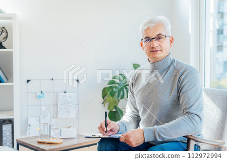 Professional Psychotherapy online. Middle aged male Psychologist having video call consultation on laptop while sitting on armchair In office. Specialist talking, counseling, helping patient online. Professional Psychotherapy online. Middle aged male Psychologist having video call consultation on laptop while sitting on armchair In office. Specialist talking, counseling, helping patient online. 112972994