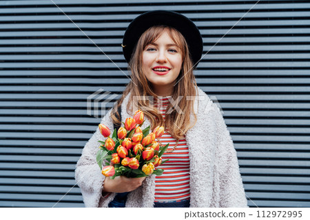 Portrait of happy, young smiling fashion woman enjoying the moment, holding bunch of fresh tulip flowers on the gray striped wall background. Urban city street fashion. Spring mood. Selective focus. 112972995