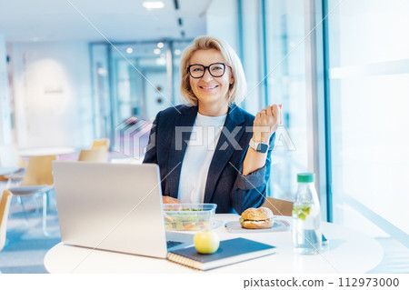 Smiling middle-aged business woman having healthy lunch at working place or business cafe, with laptop during her break. Balanced diet lunch box. Healthy eating habits and well-being. Selective focus 112973000