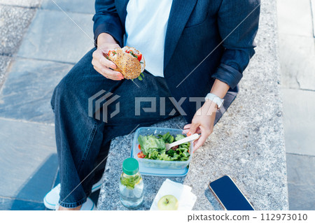 Top view close up business woman eating sandwich and fresh salad on lunch break sitting on bench at park. Balanced diet lunchbox for weight loss. Healthy eating habits and well-being. Selective focus. 112973010