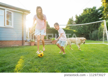 Young mother and son plays soccer with dog and have fun together. Happy family playing football with pet. Fun Playing Games in Backyard Lawn on Sunny Summer Day. Motherhood, childhood, togetherness Young mother and son plays soccer with dog and have fun together. Happy family playing football with pet. Fun Playing Games in Backyard Lawn on Sunny Summer Day. Motherhood, childhood, togetherness 112973027