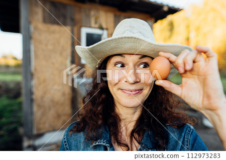 Portrait of funny joyful farmer woman holding fresh egg near her face. While enjoying farming, a female farmer holds a fresh egg Happy moment of collecting eggs Portrait of funny joyful farmer woman holding fresh egg near her face. While enjoying farming, a female farmer holds a fresh egg Happy moment of collecting eggs 112973283