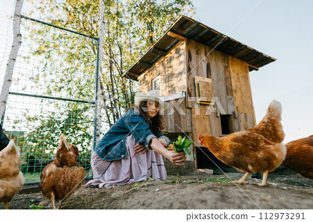 Smiling woman enjoying farm life while feeding her chickens. Happy red-haired woman enjoying feeding chickens in the garden of a rural estate Smiling woman enjoying farm life while feeding her chickens. Happy red-haired woman enjoying feeding chickens in the garden of a rural estate 112973291