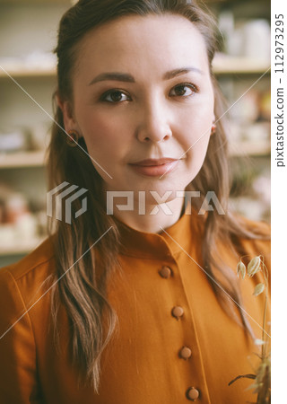 Close-up portrait of an elegant woman against the background of shelves with ceramic dishes. Elegant woman ceramic artist in a cozy studio Head shot, atmospheric portrait 112973295