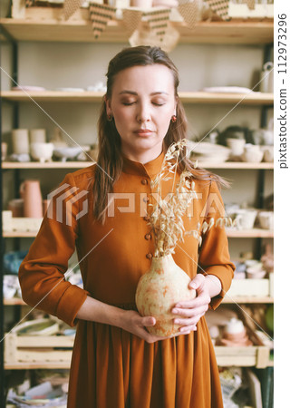 Creative Woman Potter with Clay Vase in Artisan Studio with eyes closed. Artistic woman enjoying her hobby, holding a unique piece of pottery in a well-lit studio 112973296