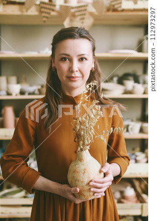 Woman Ceramic Artist Holding Artisan Clay Vase in Sunny Studio. Elegant woman holding a pottery vase with dried flowers in a sunny ceramics workshop 112973297