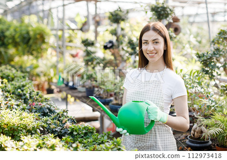 A young woman stands in the middle of a large greenhouse holding a watering can and smiling at the camera. The concept of caring for houseplants 112973418