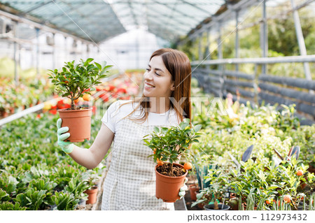 Woman holds a pot of flowers in her hands, growing plants for sale, plant as a gift, flowers in a greenhouse, potted plant. Woman holds a pot of flowers in her hands, growing plants for sale, plant as a gift, flowers in a greenhouse, potted plant. 112973432