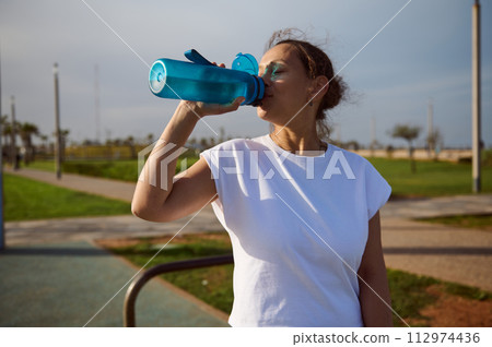 Sporty athletic woman drinking water from sports bottle while relaxing after outdoor workout on warm sunny day 112974436