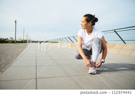 Young woman jogger tying laces on her pink sneakers, smiling looking away, getting ready for morning jog in the city Young woman jogger tying laces on her pink sneakers, smiling looking away, getting ready for morning jog in the city 112974441