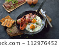 Crunchy fried eggs with Italian pancetta bacon served  in an old iron frying pan on rustic weathered wood 112976452