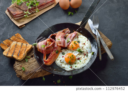 Crunchy fried eggs with Italian pancetta bacon served  in an old iron frying pan on rustic weathered wood 112976452