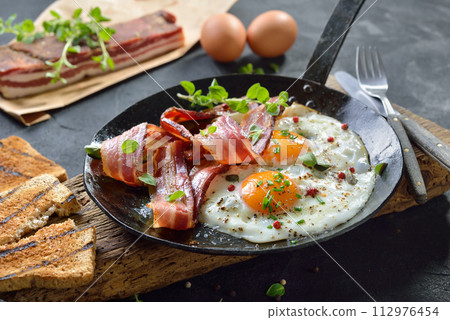 Crunchy fried eggs with Italian pancetta bacon served  in an old iron frying pan on rustic weathered wood 112976454