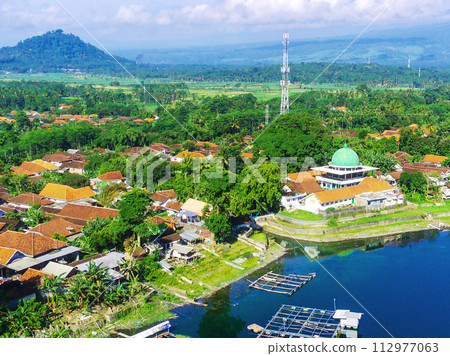 An aerial view of a small lakeside town with a mosque, surrounded by trees and houses. Fish cages are visible in the water. An aerial view of a small lakeside town with a mosque, surrounded by trees and houses. Fish cages are visible in the water. 112977063